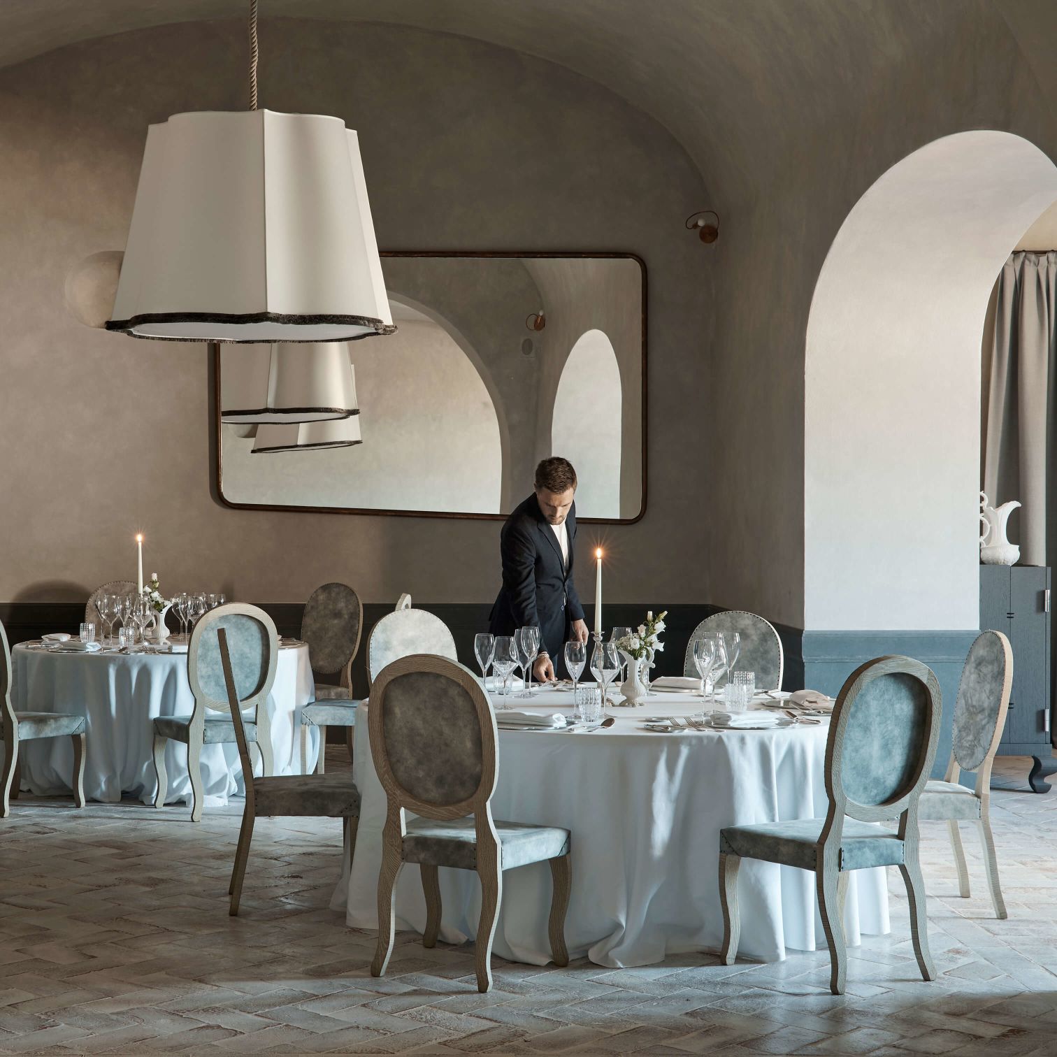 A Person Standing Next To A Table With Chairs And A Chandelier